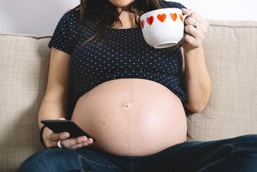 Schwangere Frau zeigt ihren Bauch, während sie mit einer Tasse Tee und einem Smartphone auf der Couch sitzt - GEMF01204