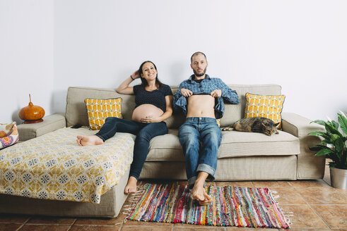 Glückliches Paar, das ein Baby erwartet, sitzt zu Hause auf der Couch mit seiner Katze - GEMF01207