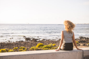 Rückenansicht einer Frau, die auf einer Mauer sitzt und auf das Meer schaut - SIPF01064
