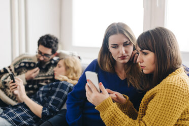 Junge Frau tauscht mit einem Freund im Wohnzimmer Nachrichten auf dem Smartphone aus - LCUF00078