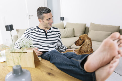 Entspannter Mann mit Hund zu Hause, der ein Tablet benutzt - MADF01183