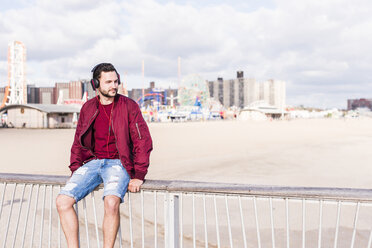 USA, New York City, Mann sitzt auf einem Geländer auf Coney Island und trägt Kopfhörer - UUF09162