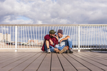 USA, New York City, two friends sitting on bridge on Coney Island sharing headphones - UUF09168