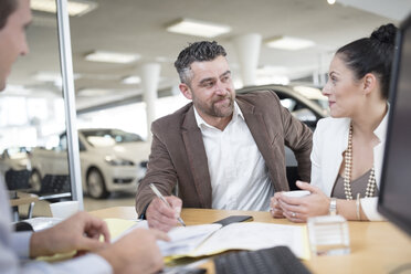 Car dealer and couple sitting at desk with agreement forms in showroom - ZEF11552