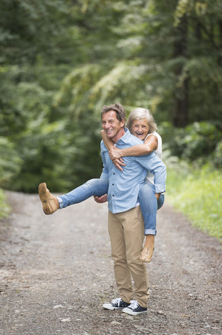 Ein älterer Mann nimmt seine Frau auf einem Waldweg huckepack, lizenzfreies Stockfoto