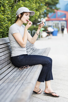 Frau sitzt auf einer Bank und benutzt einen Taschenspiegel zum Auftragen von Make-up - GIOF01598