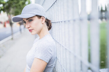 Portrait of woman wearing basecap leaning against fence - GIOF01617