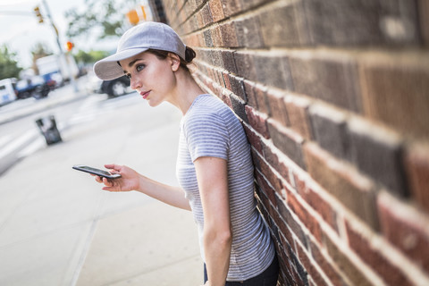 Frau mit Smartphone lehnt an einer Backsteinmauer, lizenzfreies Stockfoto