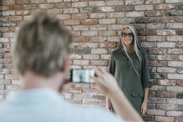 Mann, der eine lächelnde Frau mit langen grauen Haaren an einer Backsteinmauer fotografiert - KNSF00436