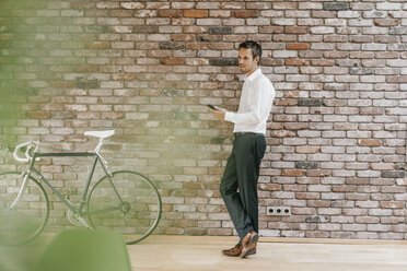 Businessman with bicycle and cell phone at brick wall - KNSF00463