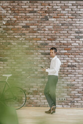 Businessman with bicycle and cell phone at brick wall - KNSF00520