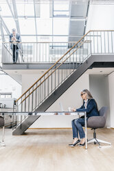 Businesswoman sitting at desk in office with businessman on upper floor - KNSF00566