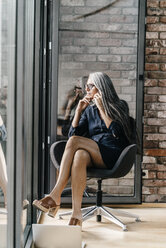 Frau mit langen grauen Haaren am Handy, am Fenster sitzend - KNSF00581