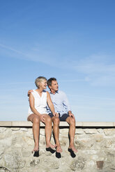 Senior couple sitting barefoot on a wall in front of sky watching something - HAPF01141