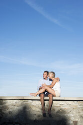 Happy senior couple sitting barefoot on a wall in front of sky - HAPF01150