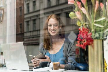 Porträt einer lächelnden Frau mit Smartphone und Laptop in einem Kaffeehaus - TAMF00808