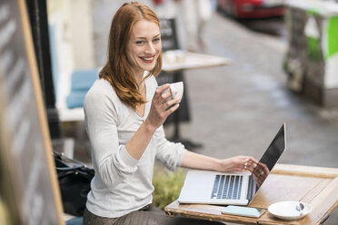 Rothaarige Frau mit einer Tasse Kaffee und einem Laptop in einem Straßencafé - TAMF00817