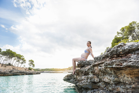 Schwangere Frau sitzt auf einem Felsen am Meer, lizenzfreies Stockfoto