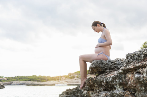 Schwangere Frau sitzt auf einem Felsen am Meer, lizenzfreies Stockfoto