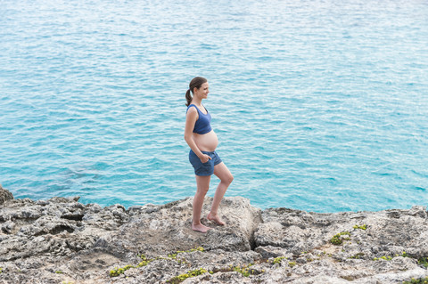 Schwangere Frau steht auf einem Felsen am Meer, lizenzfreies Stockfoto