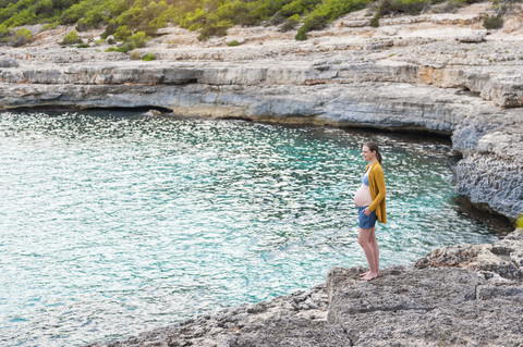 Schwangere Frau steht auf einem Felsen am Meer, lizenzfreies Stockfoto