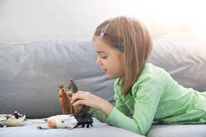 Little girl lying on couch playing with animal figurines - LVF05592