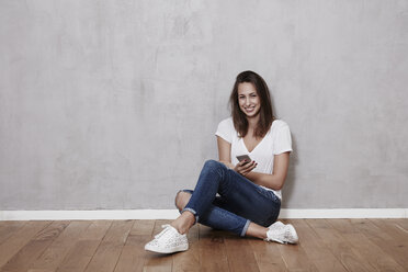 Smiling young woman sitting on the floor with cell phone - FMKF03241