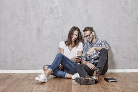 Lächelndes junges Paar sitzt mit Handy auf dem Boden, lizenzfreies Stockfoto