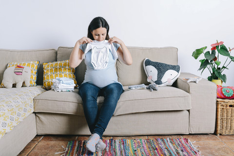 Schwangere Frau sitzt auf der Couch und sieht sich Babyschlafsäcke an, lizenzfreies Stockfoto