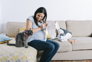 Schwangere Frau mit Katze auf der Couch sitzend, mit Blick auf Babyschlafsäcke - GEMF01241