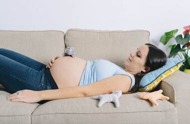 Pregnant woman lying on couch, playing with toy rhino - GEMF01248