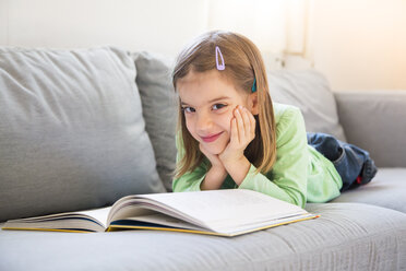 Porträt eines lächelnden kleinen Mädchens, das mit einem Buch auf der Couch liegt - LVF05606