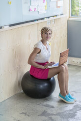 Lächelnde Geschäftsfrau, die auf einem Fitnessball sitzt und einen Laptop benutzt - RIBF00628