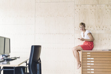 Lächelnde Geschäftsfrau, die auf einem Aktenschrank in einem kreativen Büro sitzt - RIBF00646