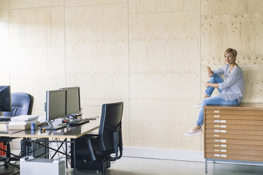 Lächelnde Geschäftsfrau, die auf einem Aktenschrank in einem kreativen Büro sitzt - RIBF00666