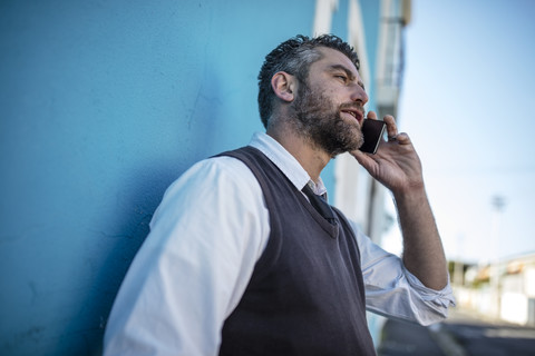 Mann lehnt an einer blauen Wand und spricht mit einem Handy, lizenzfreies Stockfoto