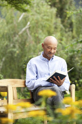 Älterer Mann sitzt auf einer Gartenbank und liest ein Buch - MFRF00751