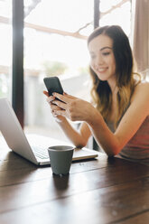 Lächelnde junge Frau benutzt Handy und Laptop zu Hause - GIOF01625