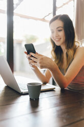 Smiling young woman using cell phone and laptop at home - GIOF01625