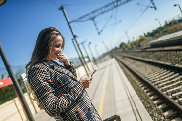 Lächelnde Frau auf dem Bahnsteig, die auf ihr Mobiltelefon schaut - KIJF00863