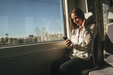 Lächelnde Frau in einem Zug, die auf ihr Mobiltelefon schaut - KIJF00865