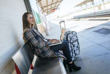 Businesswoman sitting on a bench at train station using a laptop - KIJF00895