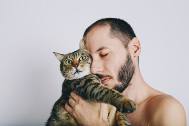 Bearded man with eyes closed holding tabby cat in front of grey background - GEMF01258