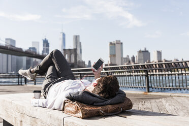 USA, Brooklyn, businesswoman lying on bench listening music with earphones - UUF09263