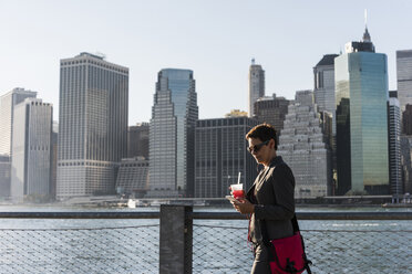 USA, Brooklyn, Geschäftsfrau schaut auf ihr Smartphone vor der Skyline von Manhattan - UUF09276