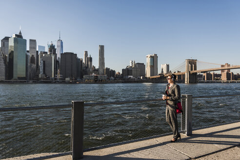 USA, Brooklyn, Geschäftsfrau mit Kopfhörern mit Blick auf die Skyline von Manhattan - UUF09277