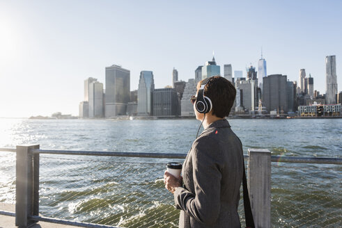 USA, Brooklyn, Geschäftsfrau mit Kopfhörern und Kaffee zum Mitnehmen vor der Skyline von Manhattan - UUF09280