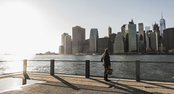 USA, Brooklyn, Rückenansicht eines Geschäftsmannes mit Aktenkoffer mit Blick auf die Skyline von Manhattan - UUF09283