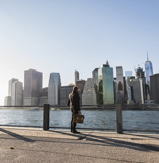 USA, Brooklyn, Rückenansicht eines Geschäftsmannes mit Aktenkoffer vor der Skyline von Manhattan - UUF09284