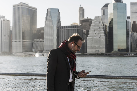 USA, Brooklyn, Geschäftsmann mit Smartphone und Kopfhörern vor der Skyline von Manhattan, lizenzfreies Stockfoto